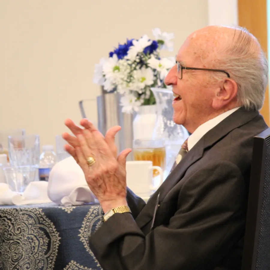 Elderly man in suit clapping hands at formal event with flower arrangement and table settings in background