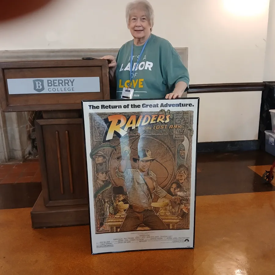 Elderly woman holding a framed Raiders of the Lost Ark movie poster beside a Berry College podium indoors.
