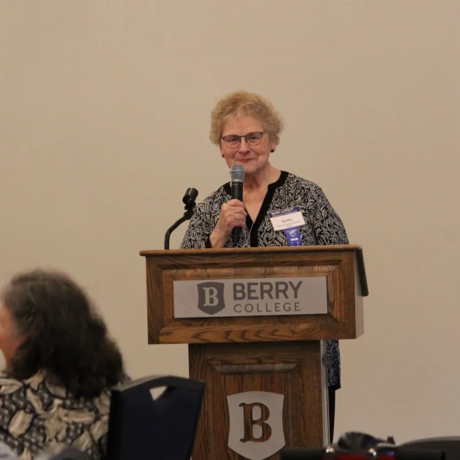 Woman speaking at Berry College podium during an indoor event with audience members seated nearby.