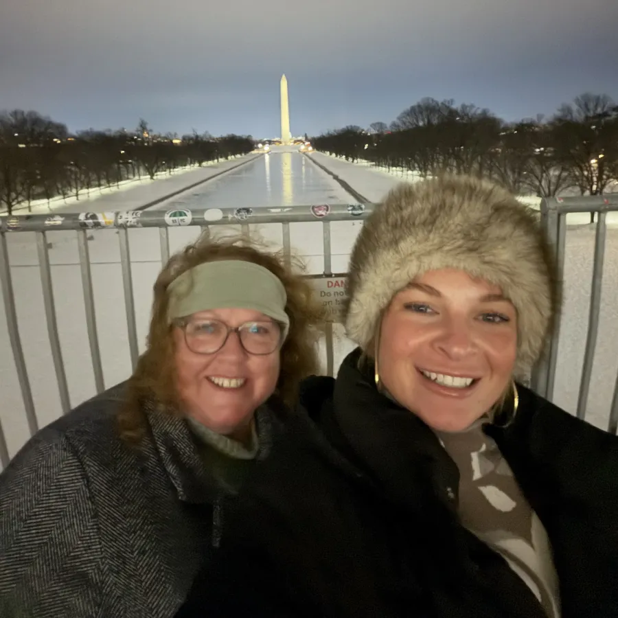 Two women smiling near a fence with the Washington Monument and reflecting pool lit up at night in the background.