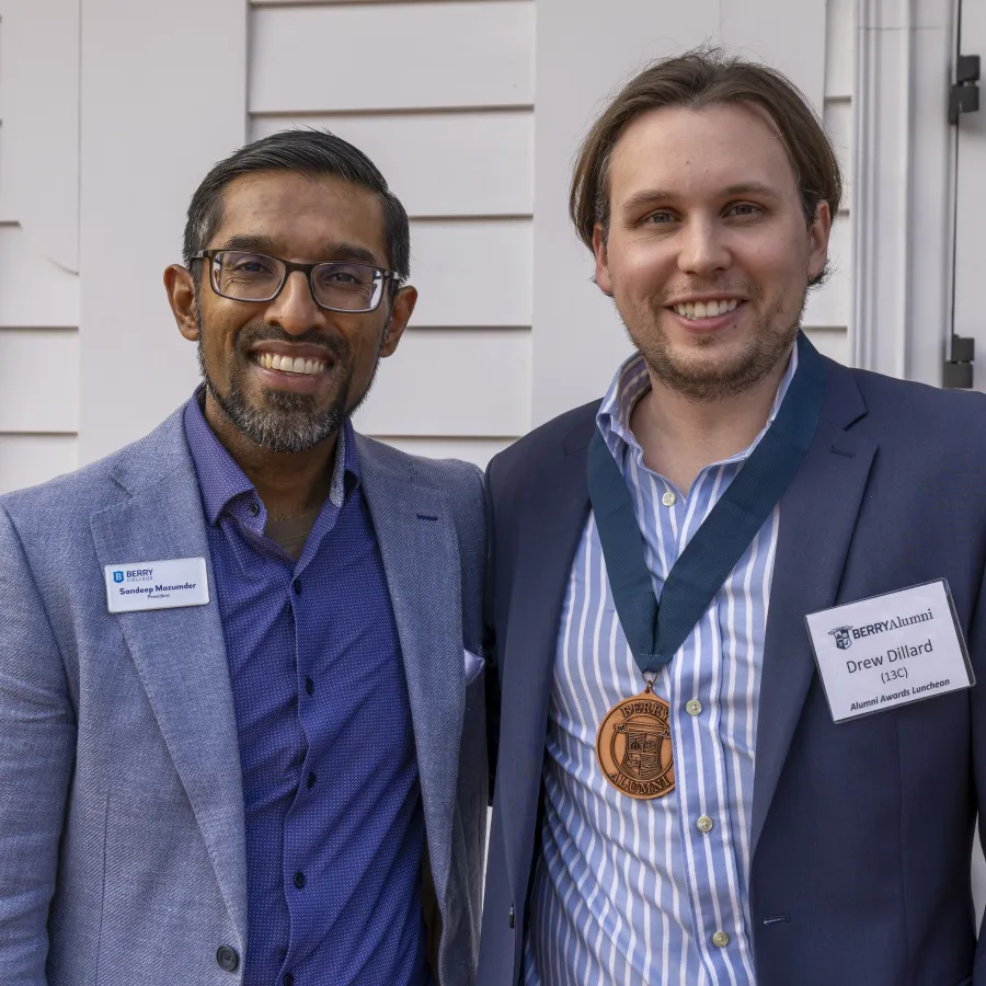 Two men in business attire smiling outside a building, one wearing a medal and both with name badges.