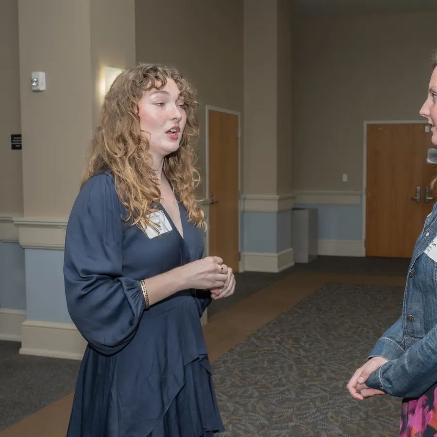 Two women engaged in conversation in a conference room with neutral walls and carpeted floor.