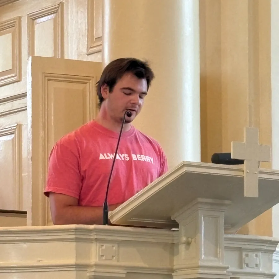 Man speaking passionately at a beige ornate podium with microphone, wearing a red shirt indoors.