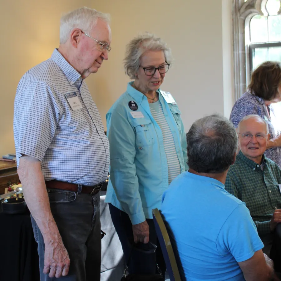 Group of elderly people engaged in conversation in a well-lit room near an arched window during a social event.