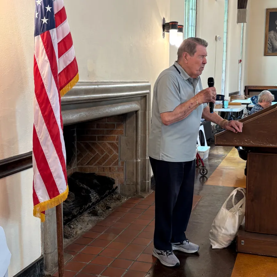 Elder man speaking into a microphone indoors with an American flag in the foreground and a fireplace in the background