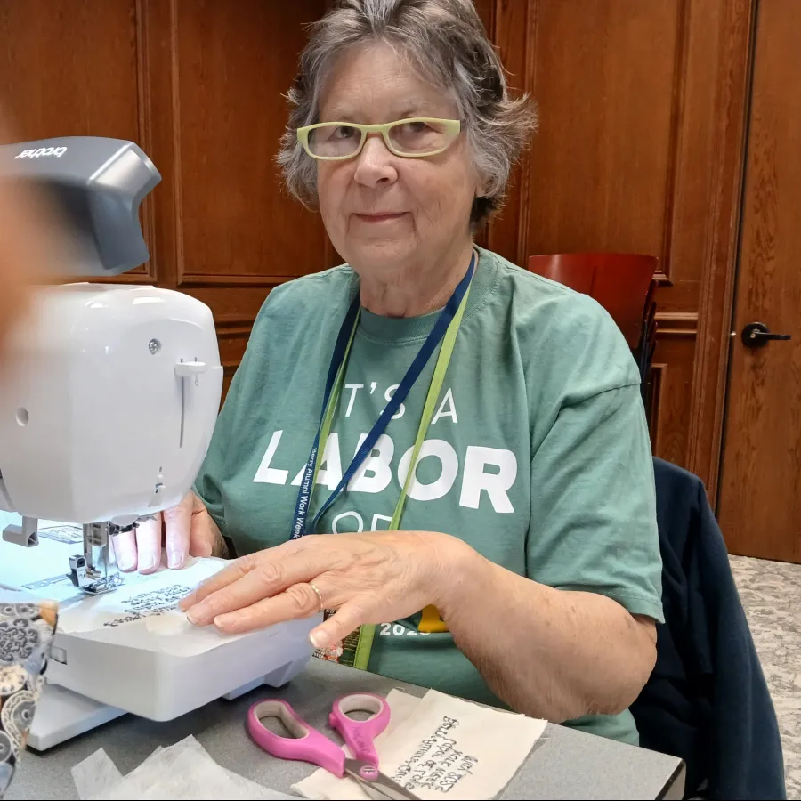 Senior woman using a sewing machine to embroider fabric with text, sitting at a table with scissors and materials.