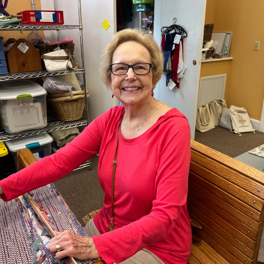Smiling elderly woman in glasses wearing a red shirt sitting at a wooden table indoors with shelves of items behind her