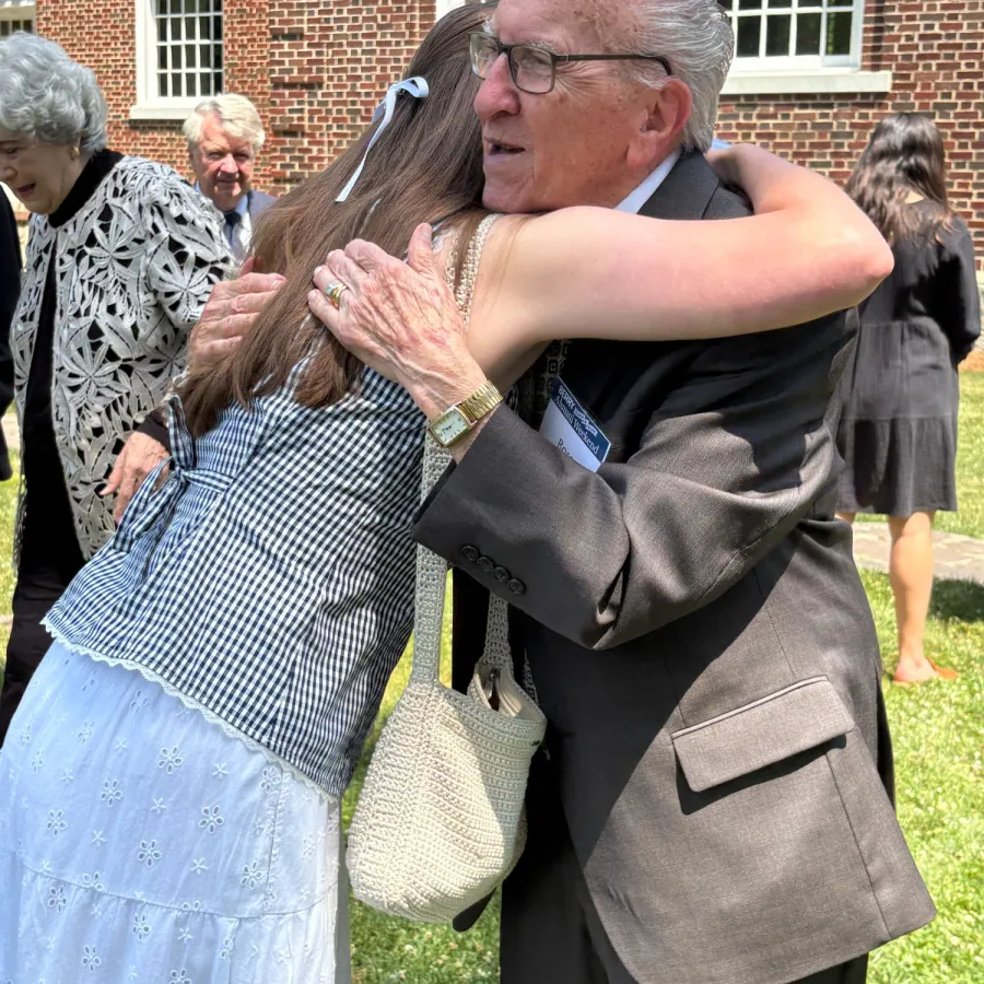 Elderly man warmly hugging a young woman outdoors near a brick building on a sunny day.