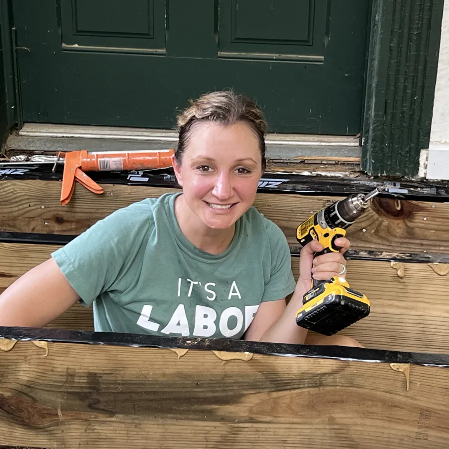 Smiling woman holding cordless drill while working on wooden porch stairs renovation project outdoors.