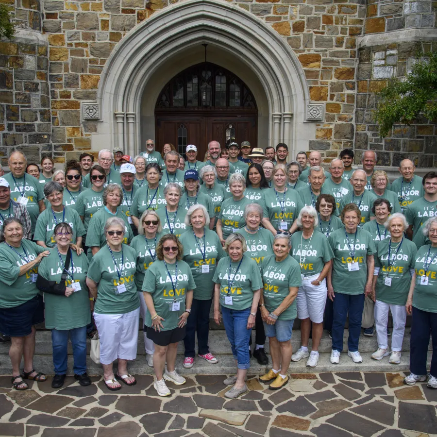Large group of adults wearing green It's a Labor of Love shirts standing in front of a stone building with arched doorway.