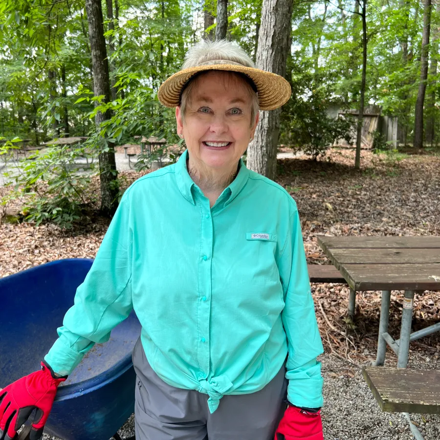 Smiling elderly woman in a blue shirt and straw hat holding a wheelbarrow in a wooded outdoor area.