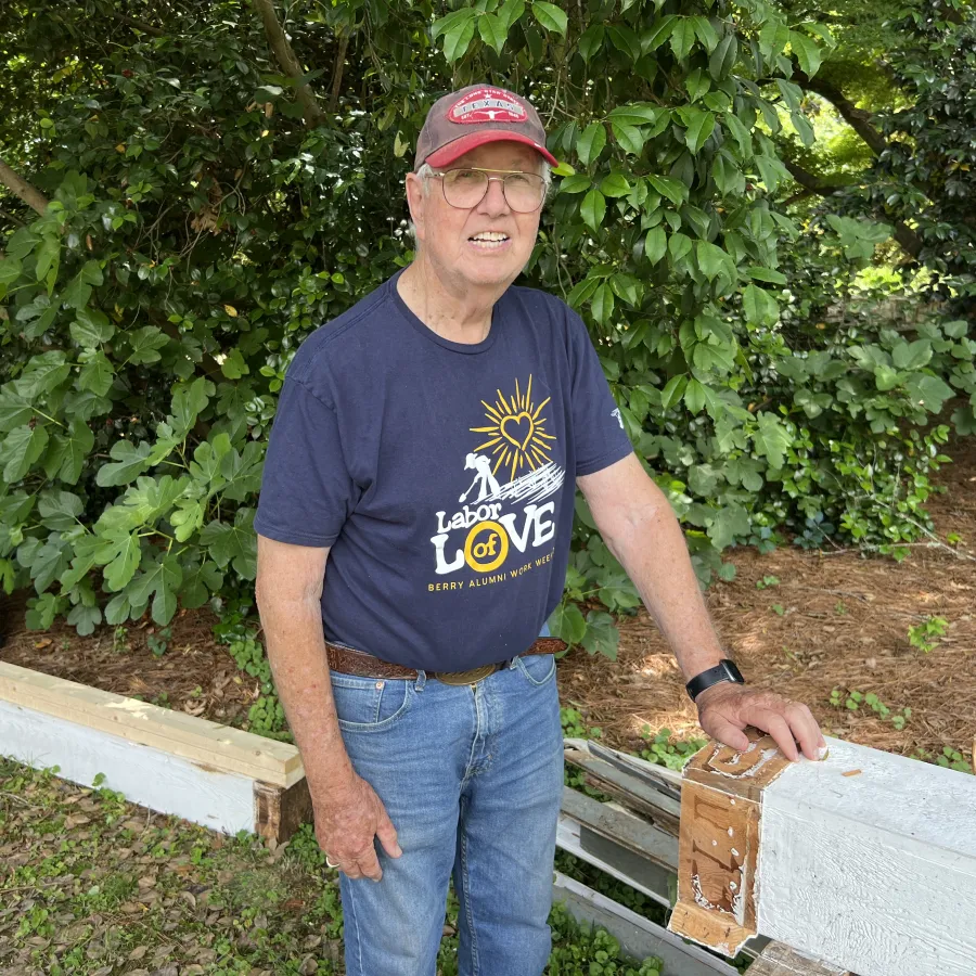 Elderly man in a red cap and Labor of Love shirt standing outdoors near wooden beams and green foliage.