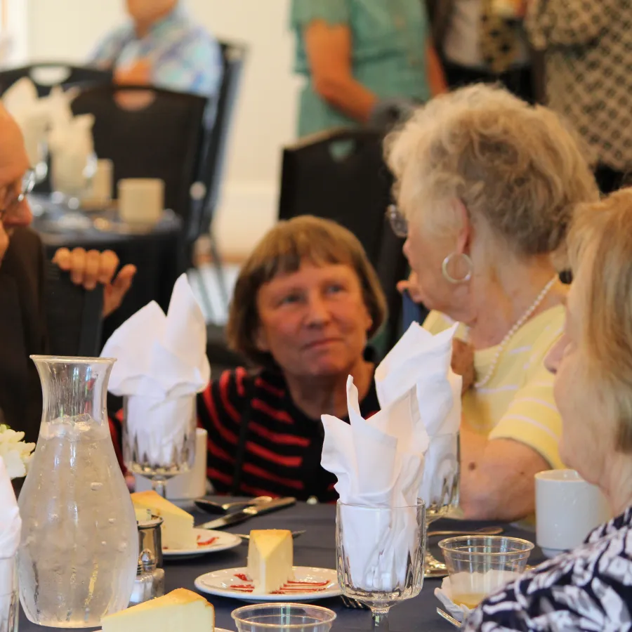 Group of elderly people engaged in conversation around a dining table set for a meal with water and cheese.