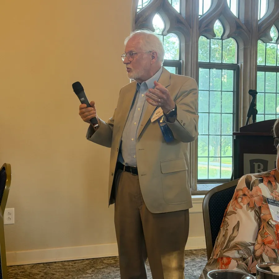 Elderly man in beige suit speaking into a microphone in a room with large arched windows and seated audience.