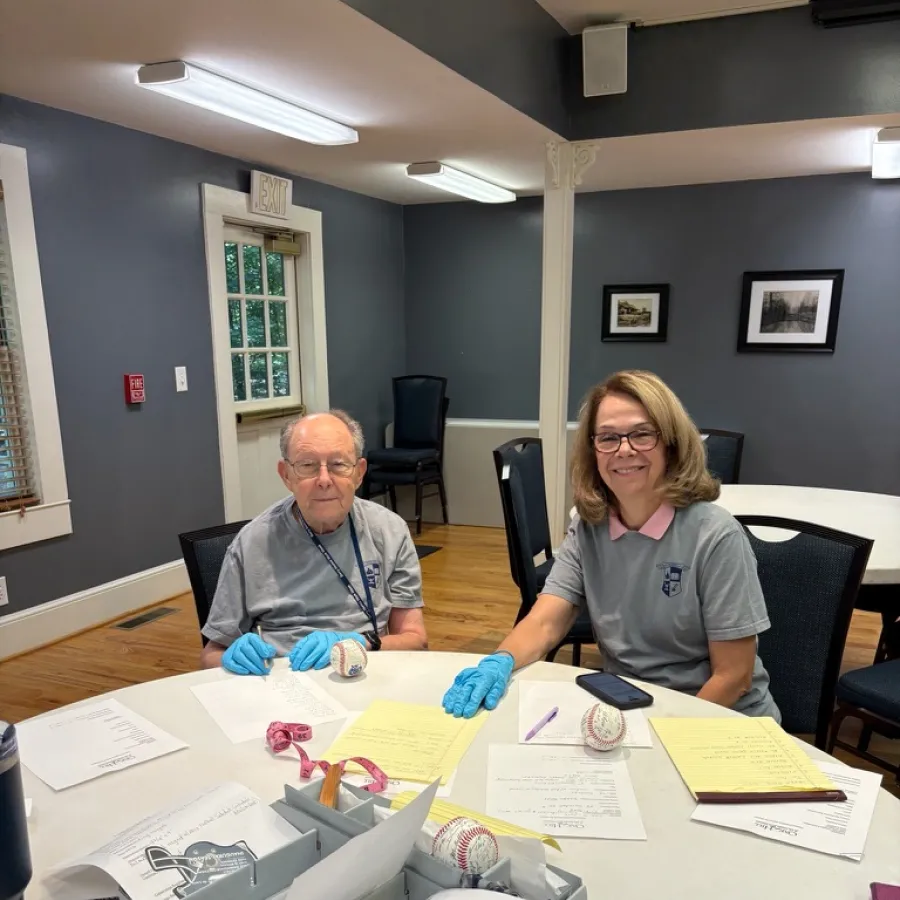 Two older adults wearing gloves sitting at a table with baseballs and paperwork in a meeting room.