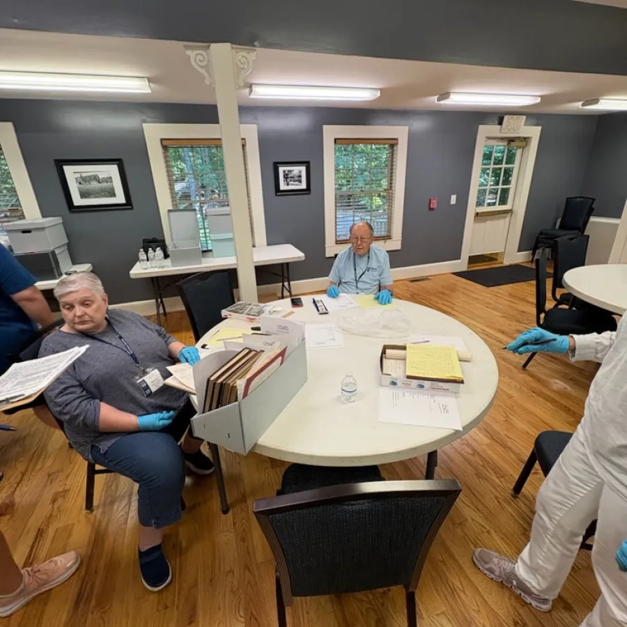People seated and standing around a table with documents in a room with wooden floors and gray walls.
