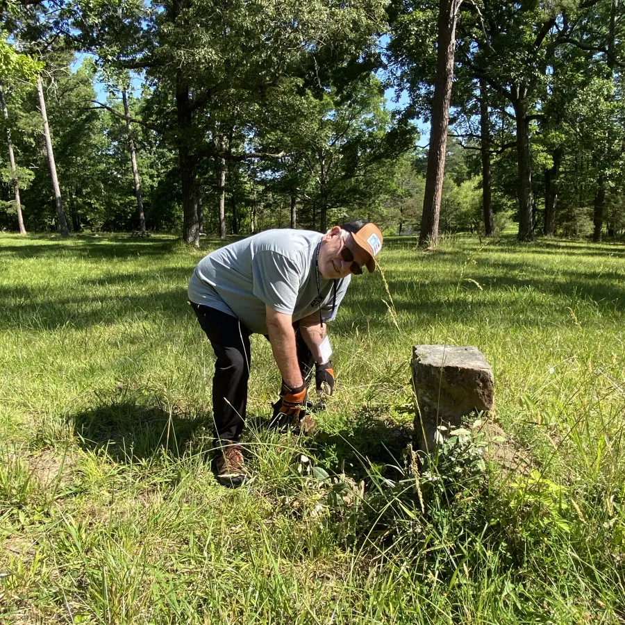 Man wearing hat and gloves clearing grass and weeds around a small stone monument in a sunny park.