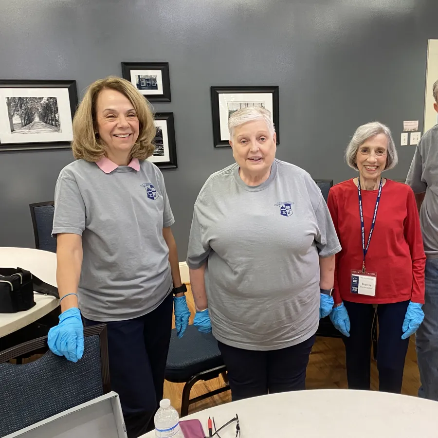 Four elderly volunteers wearing blue gloves and name tags pose indoors, three in gray shirts and one in red sweater.