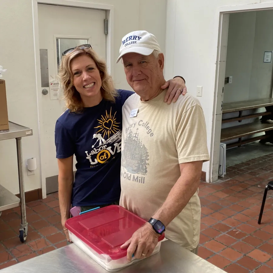 Two people smiling in a kitchen with a red container on the table and boxes nearby in a bright room.