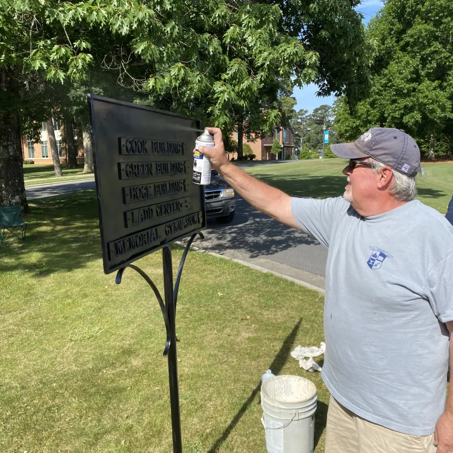 Man spray painting a black metal sign outdoors on a sunny day with another man watching nearby.