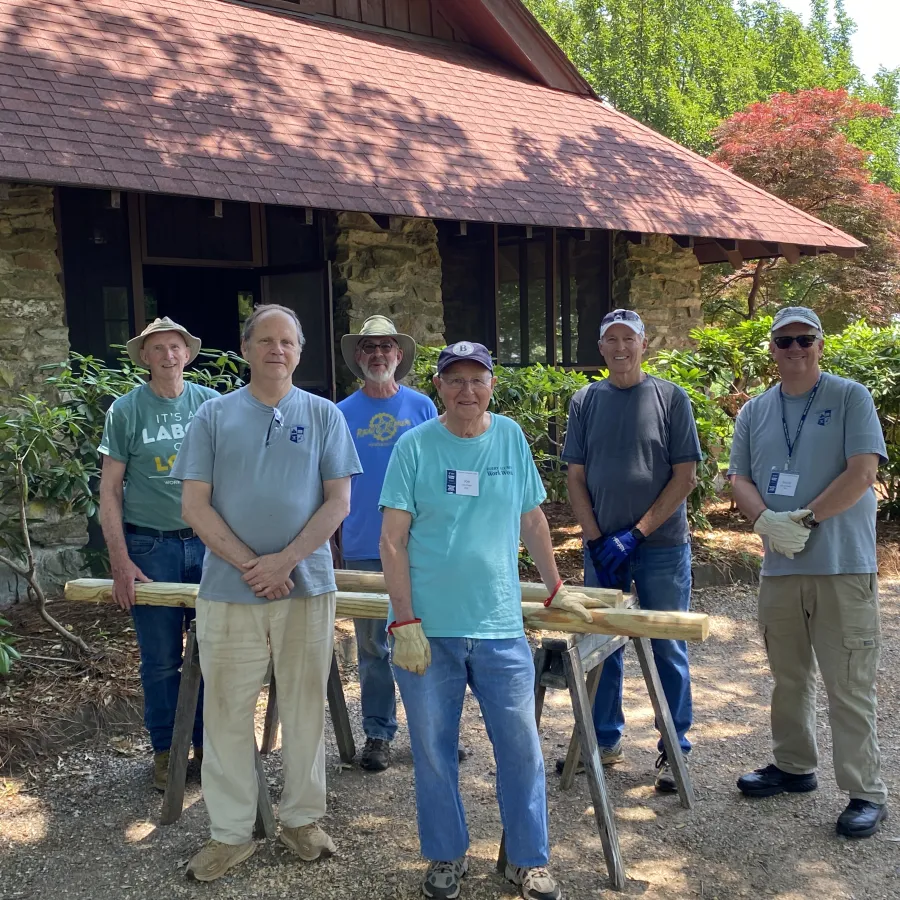 Group of six men standing outdoors with wooden logs, in front of a stone building surrounded by greenery.