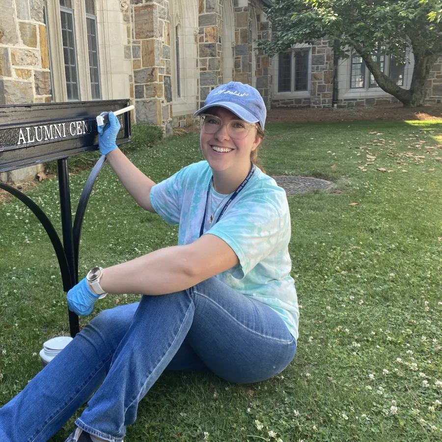 Woman in blue gloves painting black metal Alumni Center sign outdoors near stone building