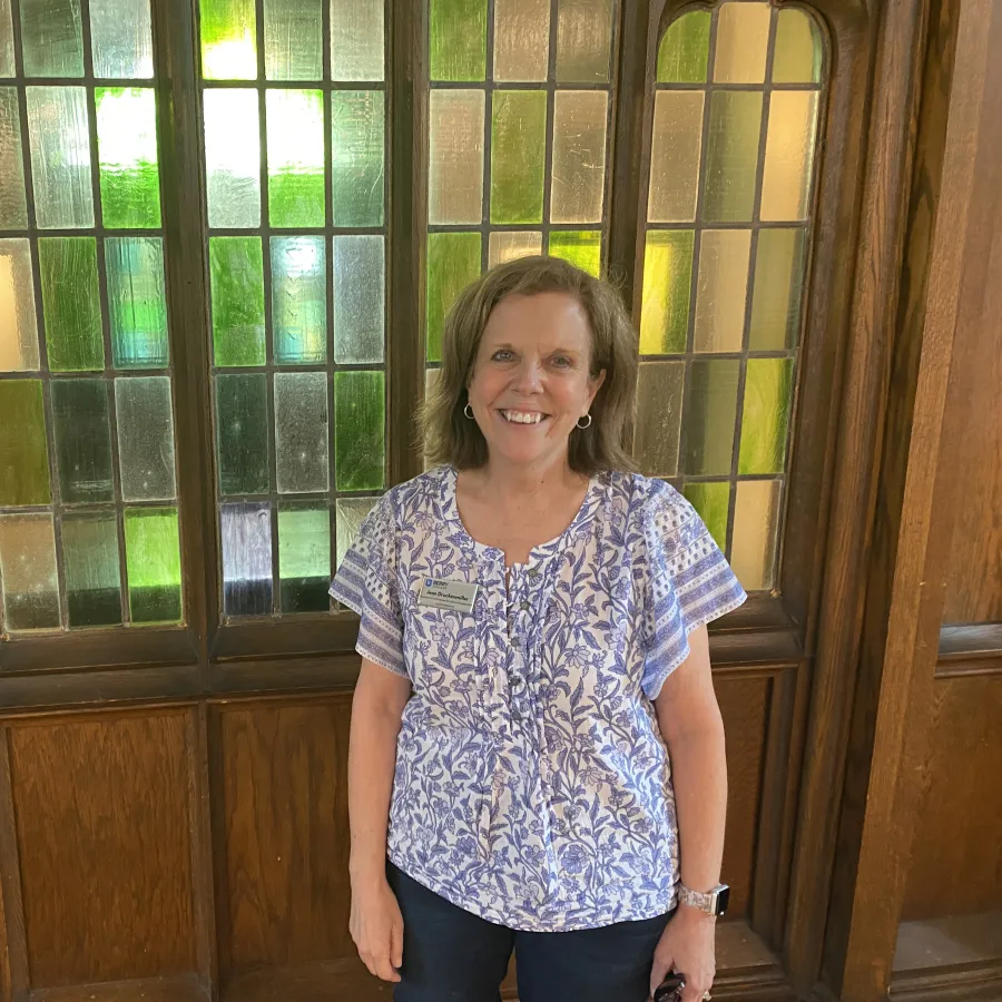 Smiling woman in floral blouse stands indoors by stained glass windows with wooden frames.