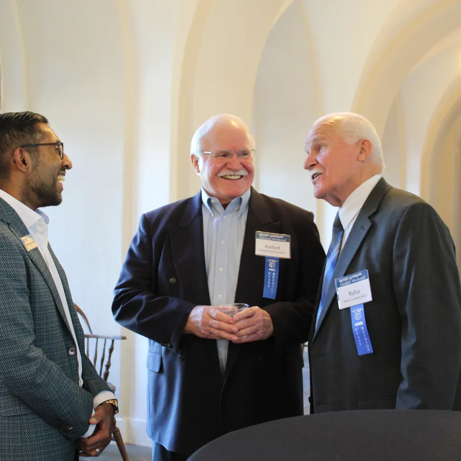 Three men wearing suits engaged in conversation at an indoor event near arched windows with name tags and ribbons.