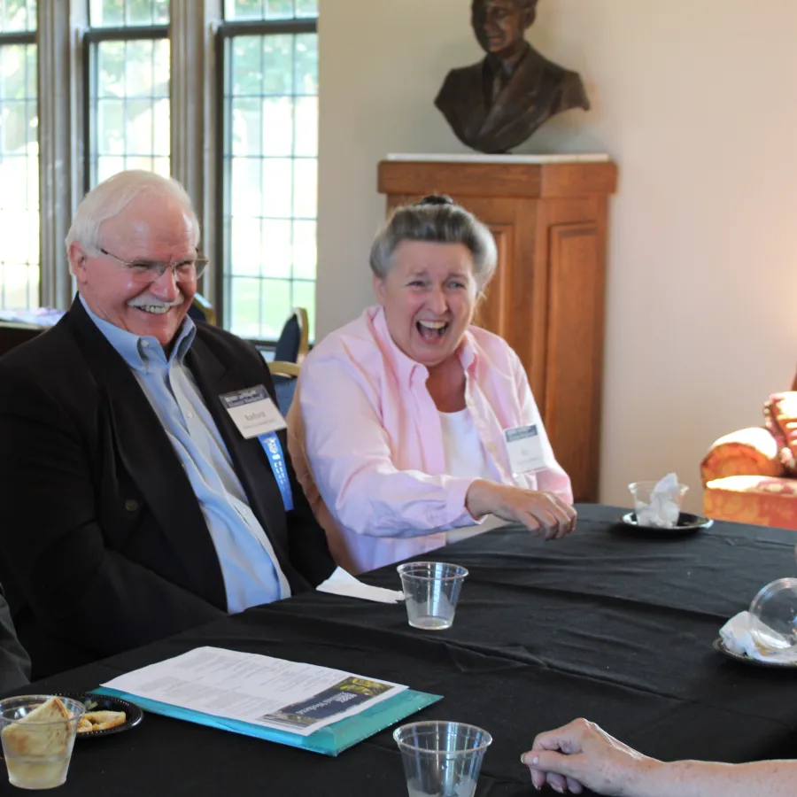 Three elderly people laughing and enjoying a conversation around a table with papers and drinks indoors