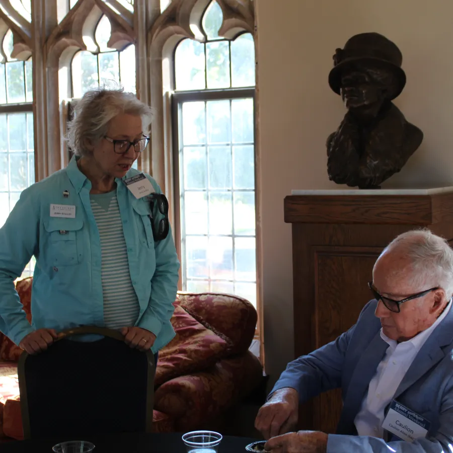 Elderly man and woman with name tags engaging in conversation in a sunlit room with gothic windows and a bust statue.
