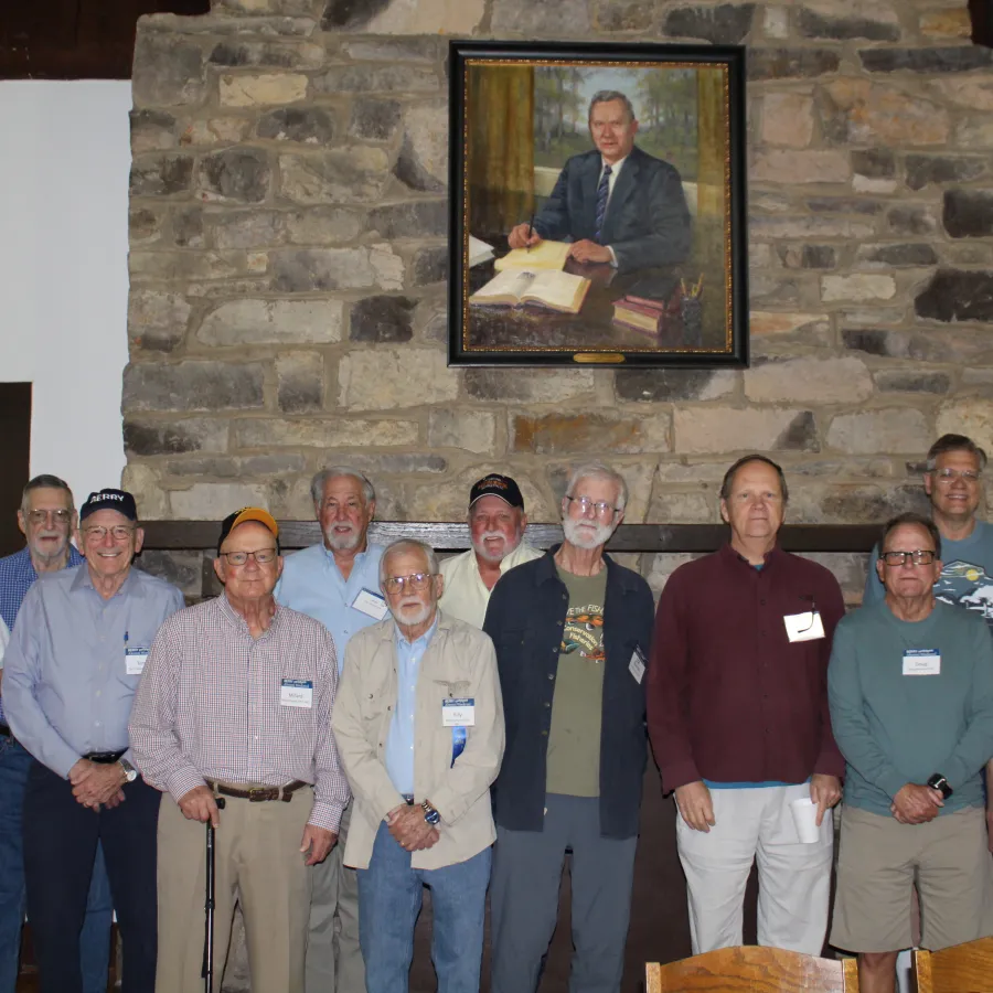 Group of older men standing indoors in front of a stone wall with a framed portrait above them.