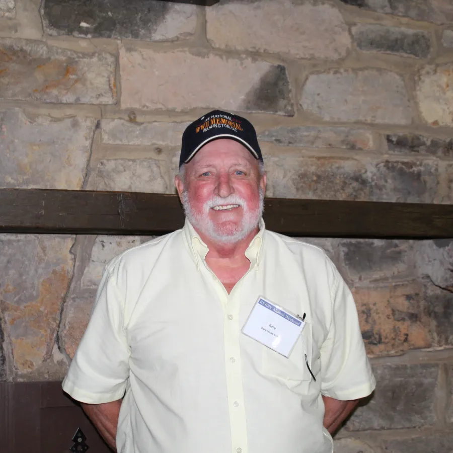Smiling elderly man with white beard wearing a light shirt, cap, and name tag stands in front of stone wall indoors.