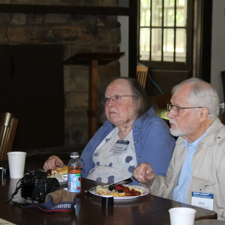 Elderly man and woman seated at a table eating a meal with camera and drinks, indoors at a social event