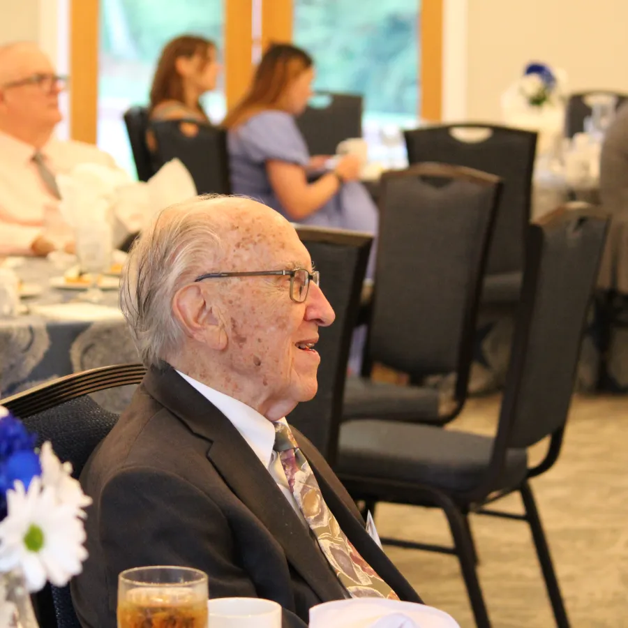 Elderly man in suit smiling and seated at a formal event with blue and white flowers on the table.