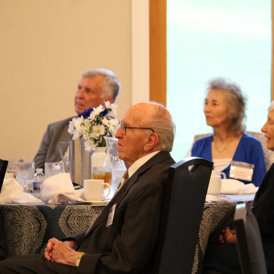 Group of elderly people seated at a table during a social or formal gathering inside a bright room.