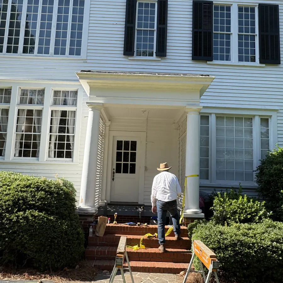 Man in hat working on brick steps at entrance of white house with columns and black shutters