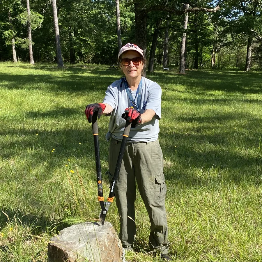Person wearing gloves and cap using loppers to cut a branch outdoors in a sunny grassy area with trees.