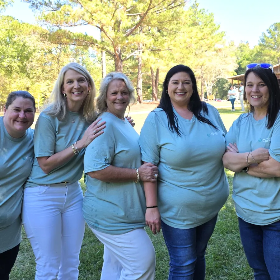 Five women in matching light blue t-shirts pose outdoors on a sunny day with trees and grass in the background.