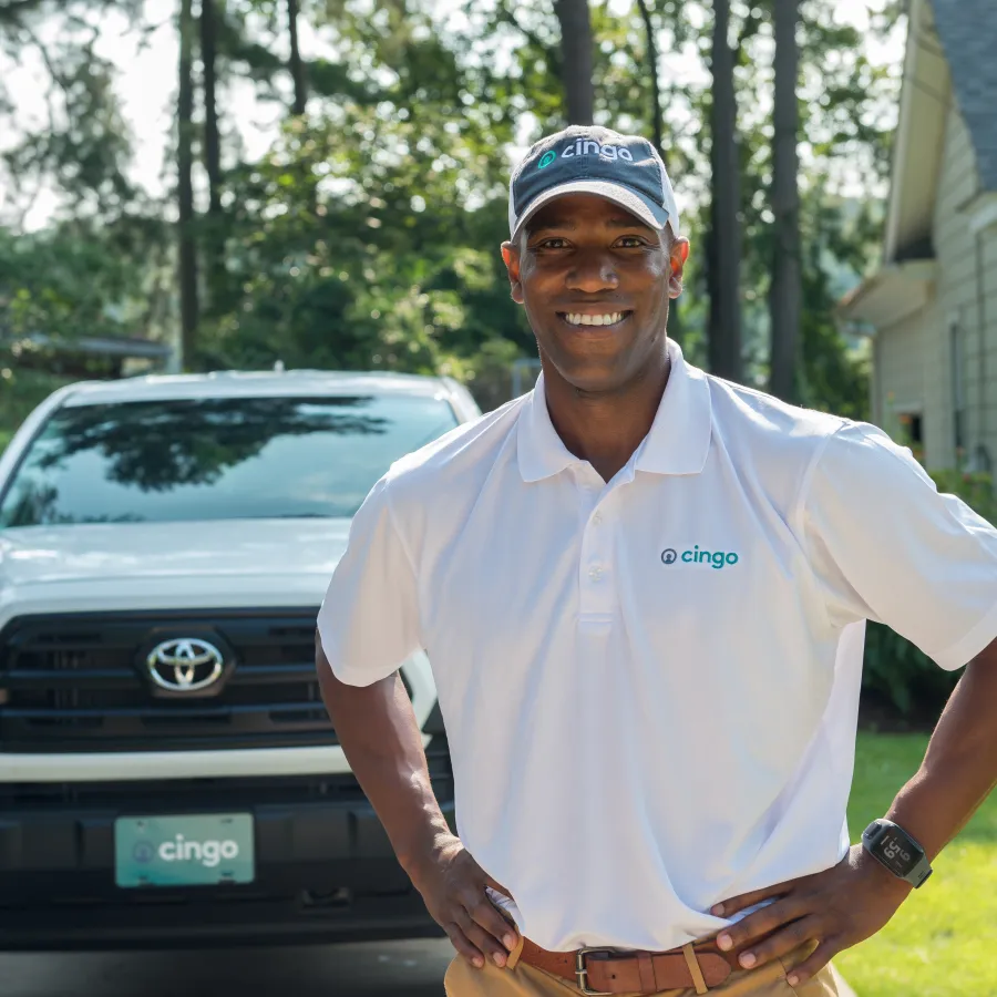 Smiling technician in Cingo uniform standing in front of a white Toyota truck in a residential driveway.