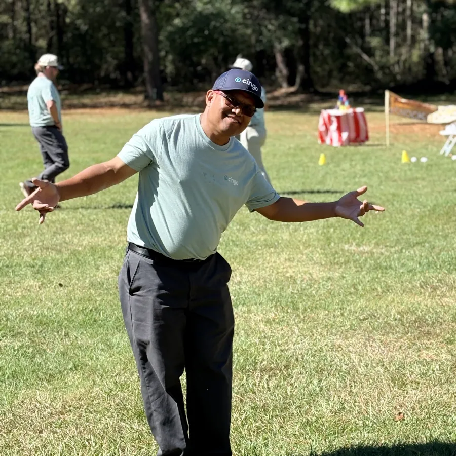 Man in gray shirt and cap posing with arms outstretched on grassy field during sunny outdoor event.