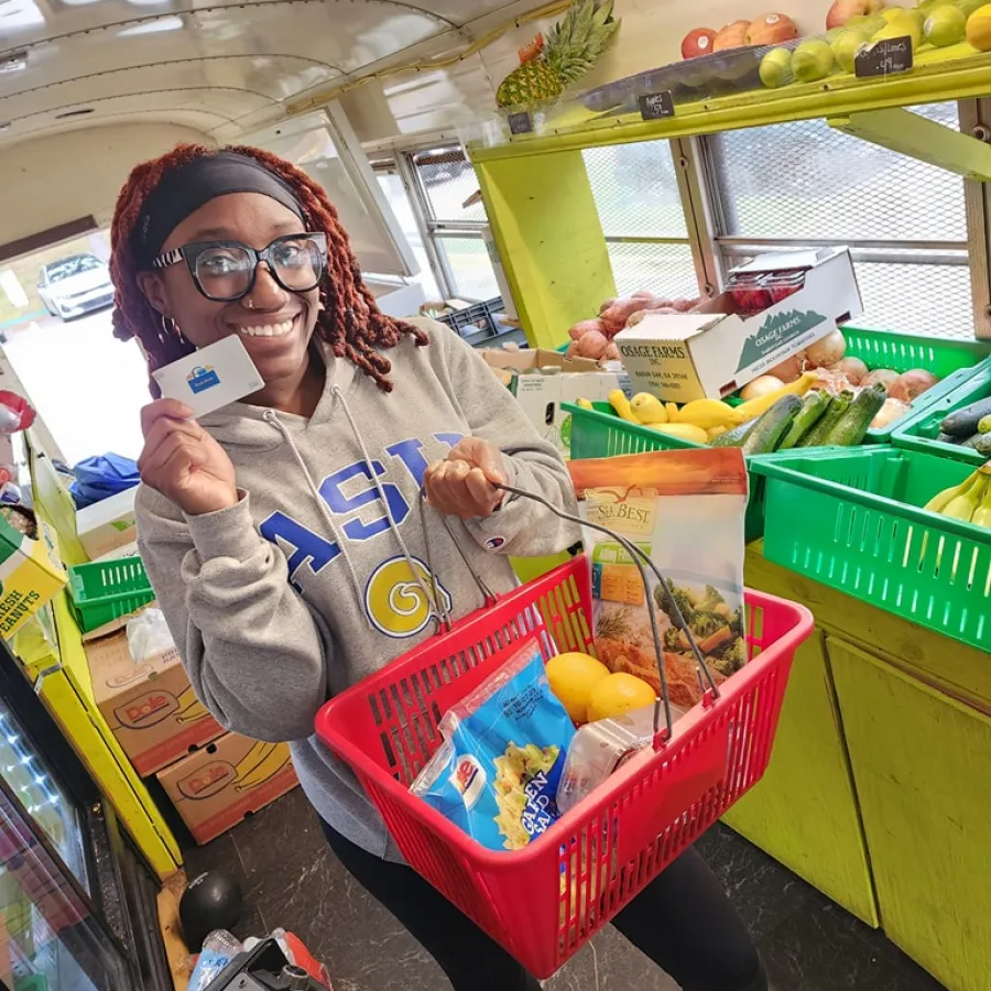 Smiling woman holding a shopping basket with groceries and a card inside a colorful fresh produce store.