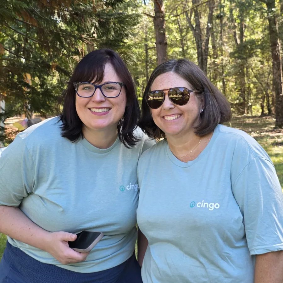 Two women wearing matching light blue Cingo t-shirts smiling outdoors in a sunlit wooded area.
