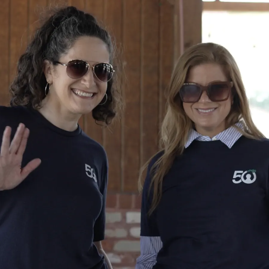 Two smiling women wearing sunglasses and navy 50th anniversary t-shirts waving indoors at a casual event