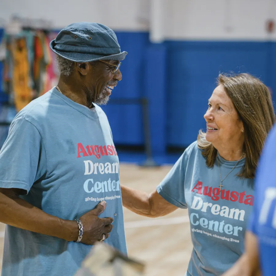 Two smiling adults wearing Augusta Dream Center shirts sharing a warm moment indoors at a community event.
