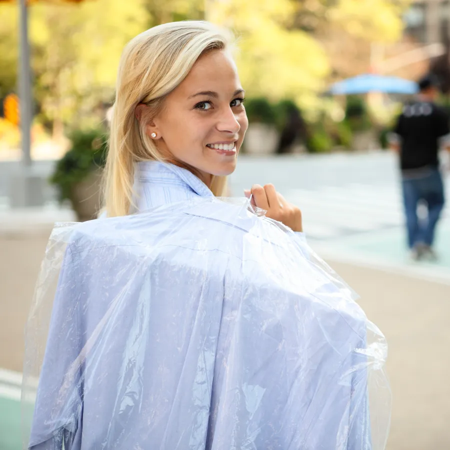 Smiling blonde woman carrying freshly dry-cleaned clothes in a plastic cover on city sidewalk.