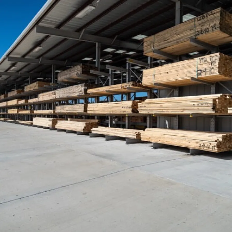 Stacks of lumber organized on metal racks under a large roof inside a spacious outdoor lumber yard.