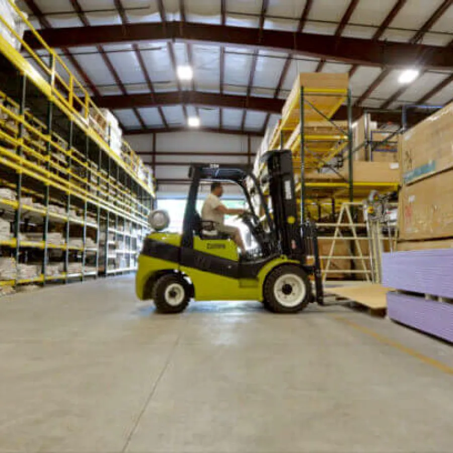 Worker operating a forklift in a warehouse moving stacked drywall sheets alongside storage racks.