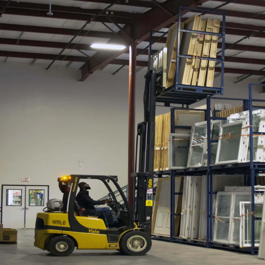 Man operating yellow Yale forklift lifting wooden frames in a warehouse with metal shelving and packaged windows.