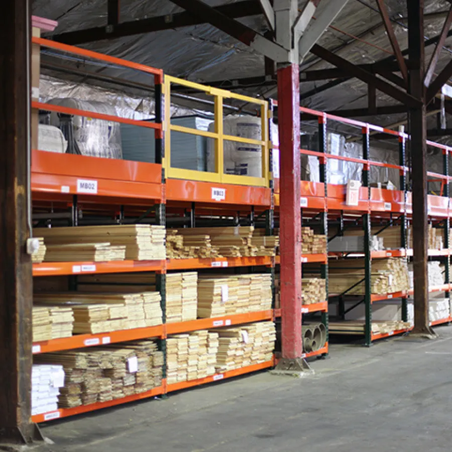 Warehouse shelves stocked with stacked wooden planks and materials under a high ceiling with metal beams.