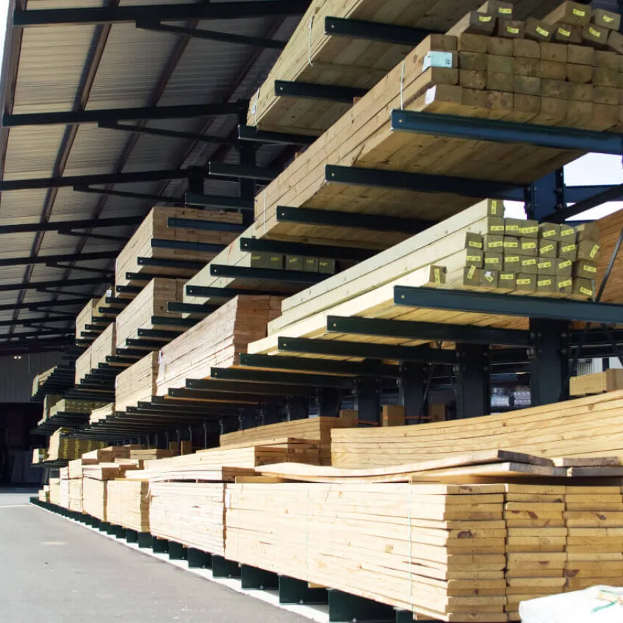 Stacks of various sizes of wooden planks stored on metal racks in an outdoor lumberyard with a partially covered roof.